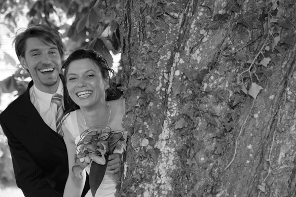 brautpaar schaut gemeinsam hinter einem dicken baum hervor, lachend, schwarz-weiss